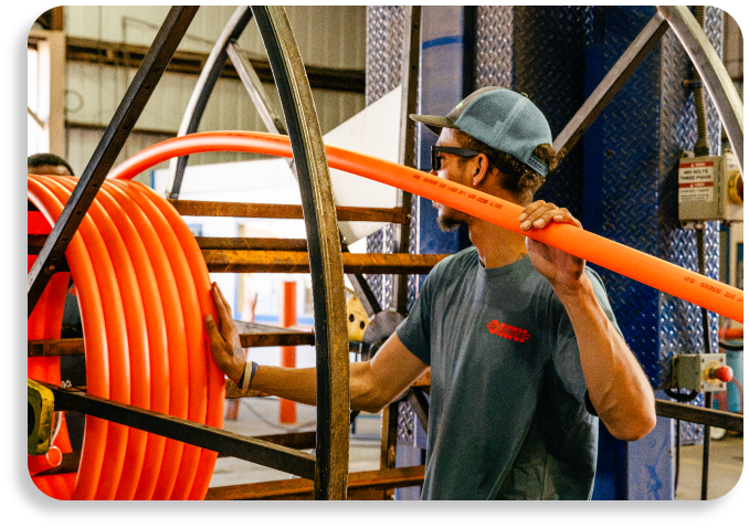 A factory specialist wearing a hard hat guiding an orange continuous HDPE conduit line off a heavy industrial metal reel.