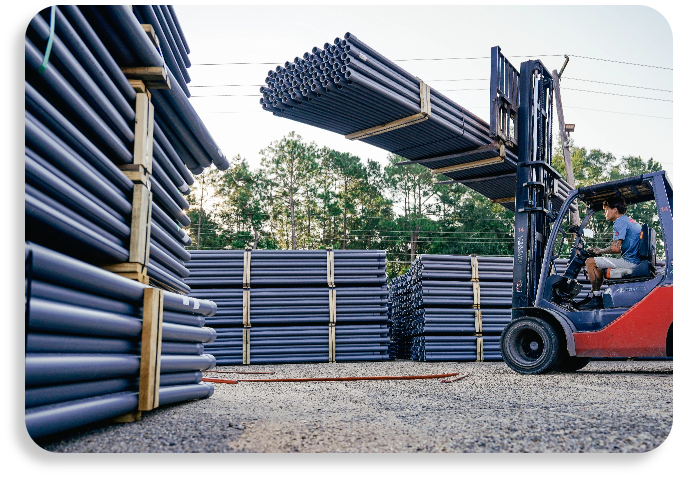 An industrial forklift operator maneuvering large stacked bundles of dark PVC pipes across factory yard logistics grounds.