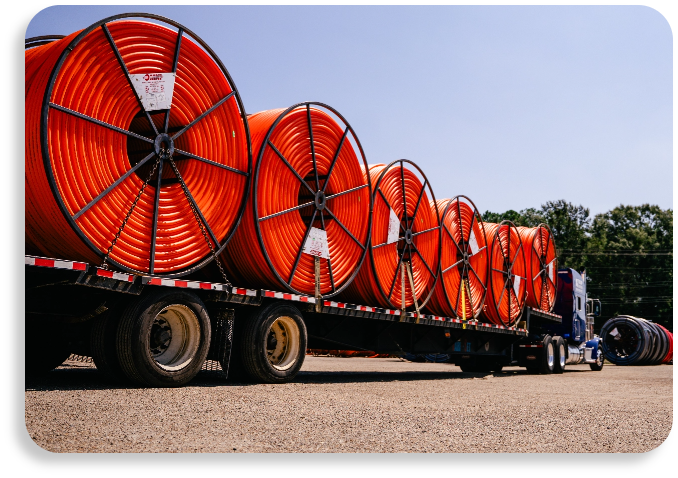 A flatbed commercial shipping truck carrying massive orange reels of continuous HDPE conduit lines outdoors.